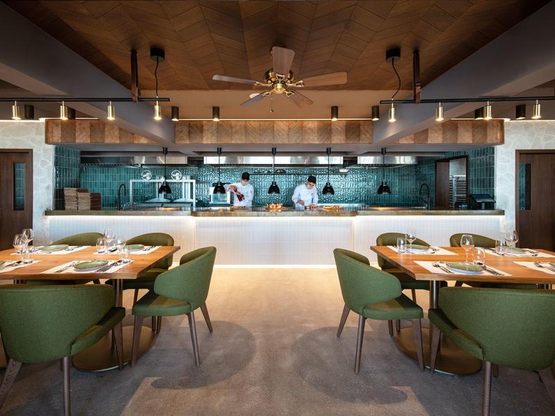 Interno di ristorante moderno con sedie verdi, tavoli apparecchiati e cucina a vista.