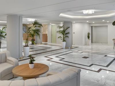 Prostorná hotelová hala s okružní pohovkou, dřevěným stolkem a dekorativními rostlinami.