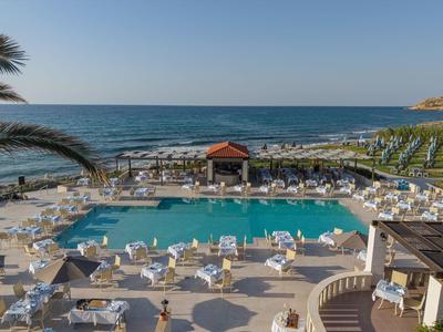 Großer Hotelpool mit Liegestühlen, Sonnenschirmen und Blick auf das offene Meer bei klarem Himmel.