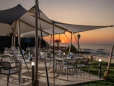 Terrasse mit mehreren Tischen und Stühlen unter Sonnensegeln mit Blick auf den Sonnenuntergang am Meer.
