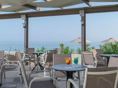 Offene Terrasse mit Tischen und Stühlen, Blick auf Meer und blauen Himmel im Hintergrund.