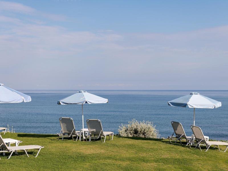 Lehátka s bílými slunečníky na trávníku u klidného moře pod jasnou oblohou.
