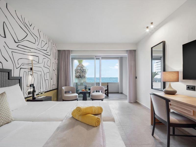 Moderní hotelový pokoj s manželskou postelí, psacím stolem, křesly a výhledem na moře z okna.