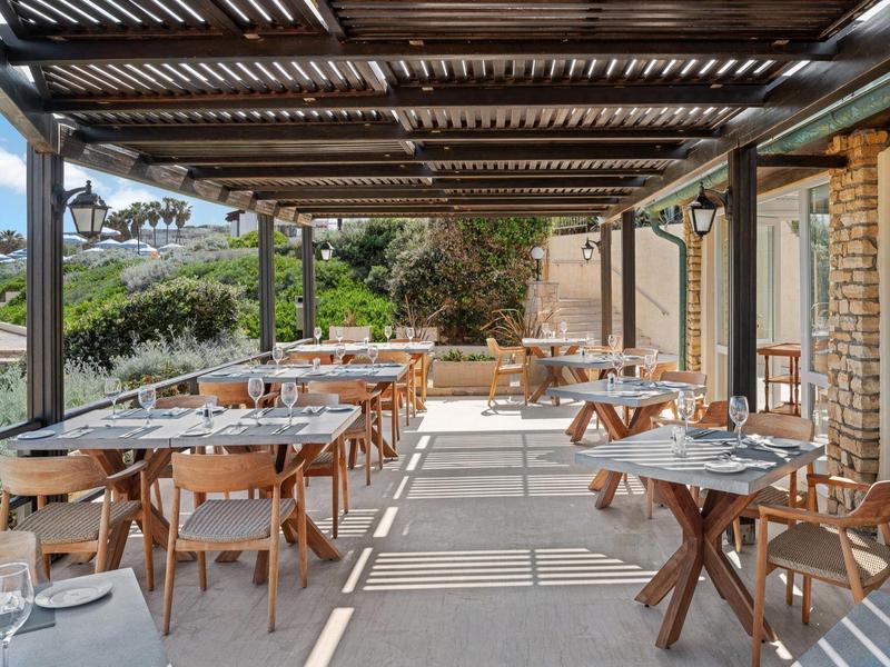 Restauranční terasa s dřevěnými stoly a židlemi pod dřevěnou pergolou.