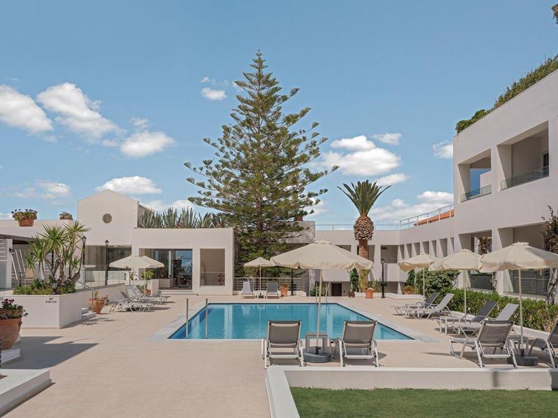 Moderní hotelové nádvoří s bazénem, lehátky, palmami a jasně modrou oblohou.