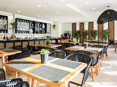 Helle, moderne Cafeteria mit Holz- und Metallmöbeln, großen Fenstern und vielen grünen Pflanzen.