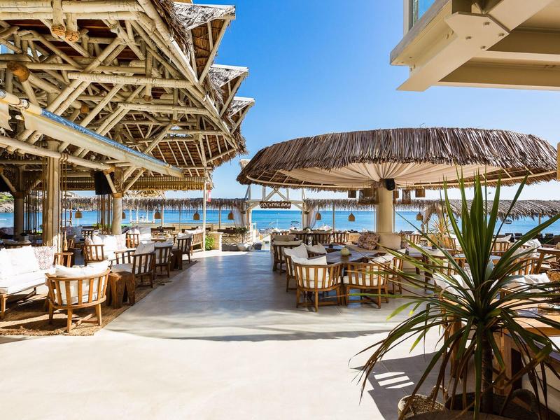 Open-air restaurant with wicker chairs and large sun umbrellas by the sea under a blue sky.