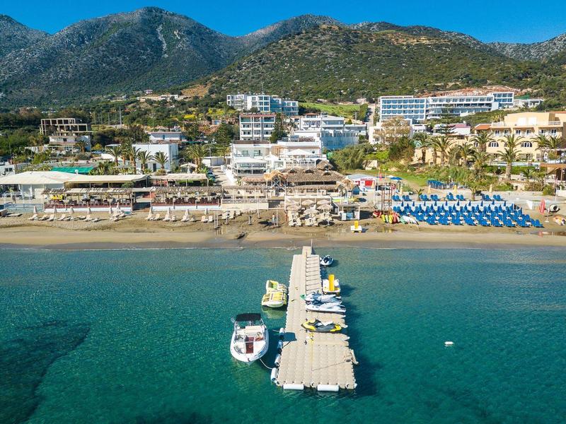 Clear blue ocean with small pier, beach, and hotels in front of forested mountains.
