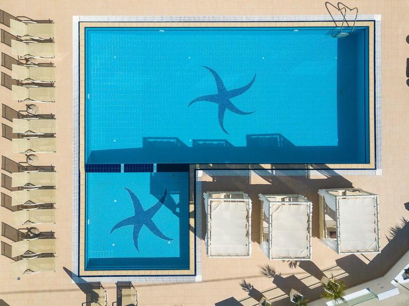 L-shaped blue pool with white lounge chairs and umbrella shadows on light flooring.