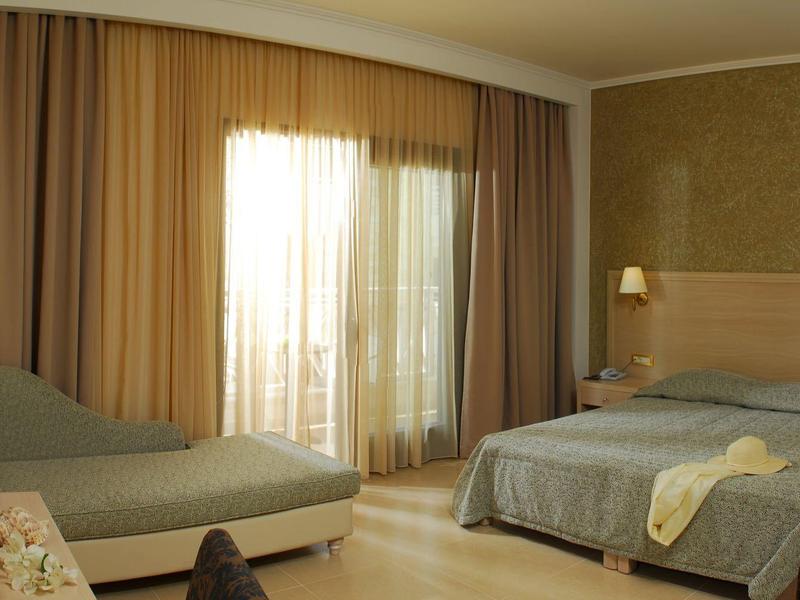 Habitación de hotel con cama doble, sofá, cortinas beige y luz natural entrando por la ventana.