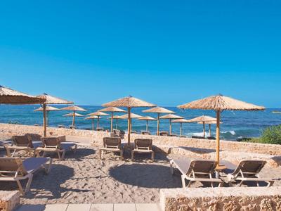 Strand mit Sand, Liegestühlen und Sonnenschirmen vor ruhigem blauem Meer und klarem Himmel.