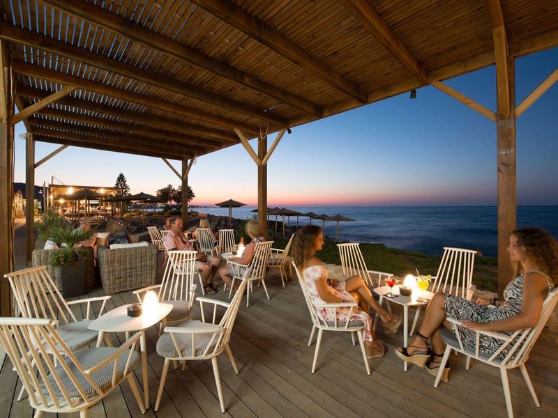 Terrazza serale con ospiti, sedie e candele sulla costa al tramonto.