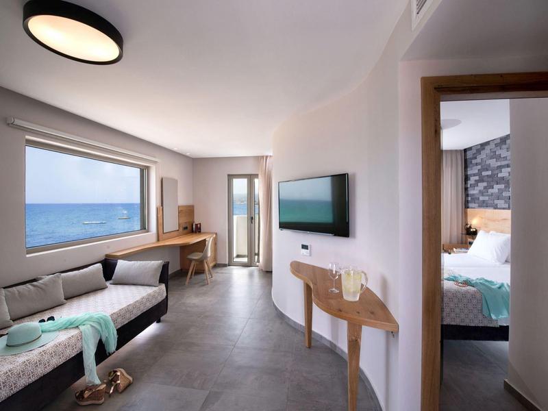 Camera d'hotel moderna con vista mare, divano, TV e camera da letto adiacente.