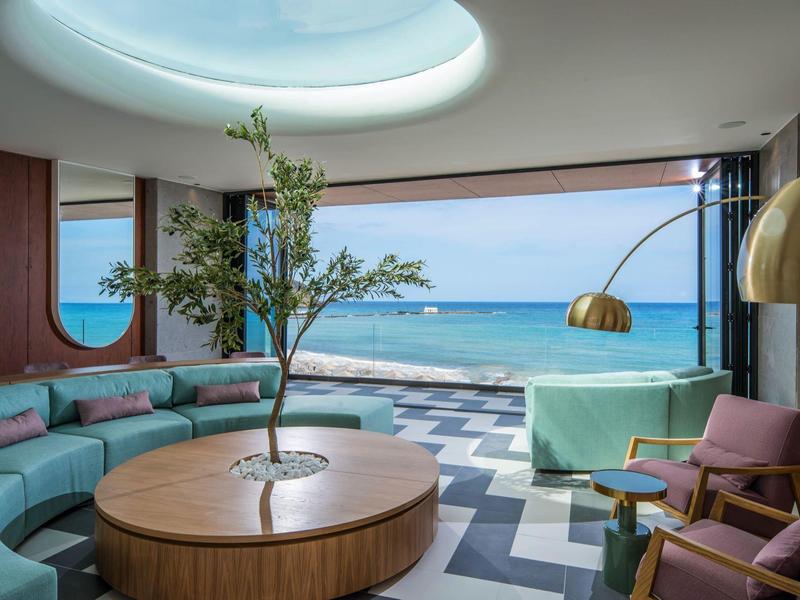 Modern lounge room with sea view, round wooden table with tree, and blue upholstered seating.