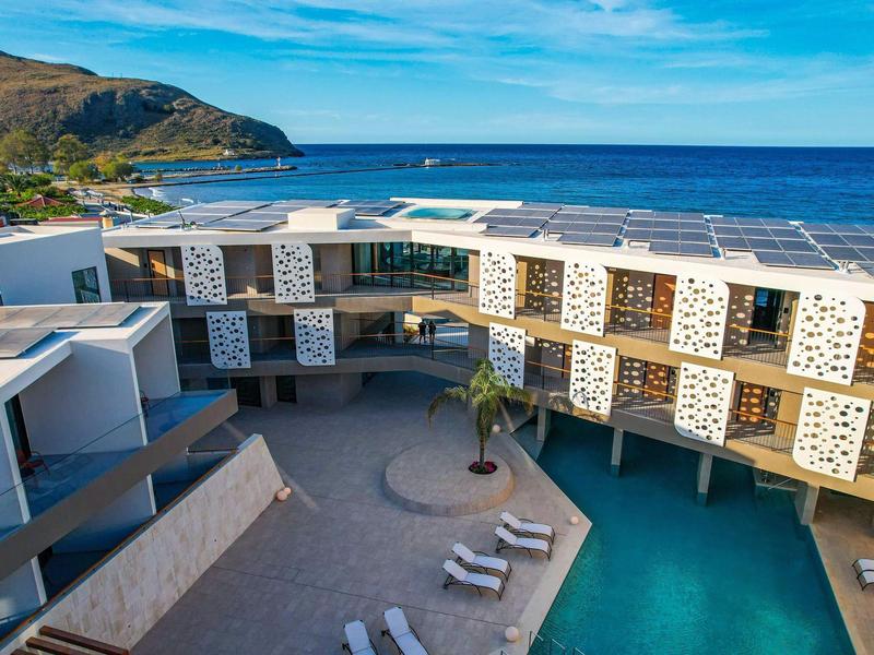 Modern seaside hotel with swimming pool and lounge areas, surrounded by mountains.