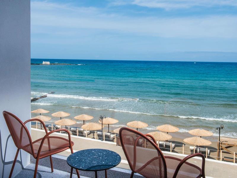 Balcony with chairs and table overlooking a sandy beach with umbrellas by the sea.