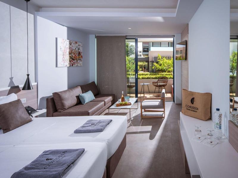 Modern hotel room with two beds, sofa, and balcony overlooking garden.