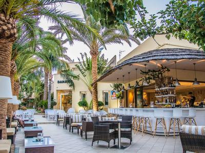 Venkovní restaurace s bílými stoly, židlemi a barem pod palmami a osvětlením.