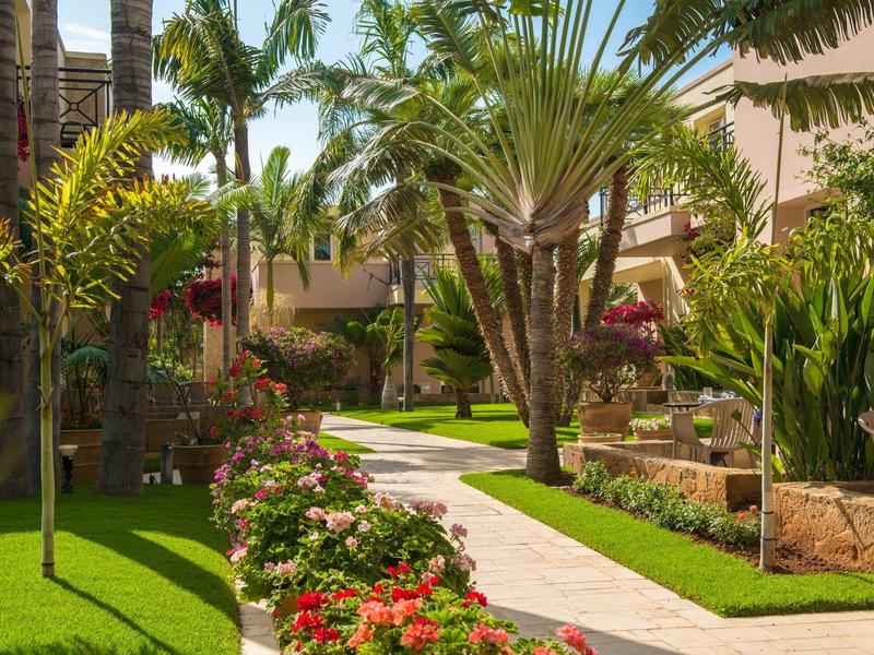 Udržovaná zahradní cesta v tropickém hotelovém areálu s palmami a barevnými květinami.