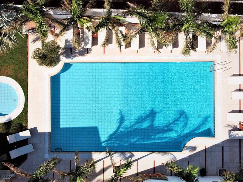 Letecký pohled na obdélníkový bazén s palmami a lehátky venku.