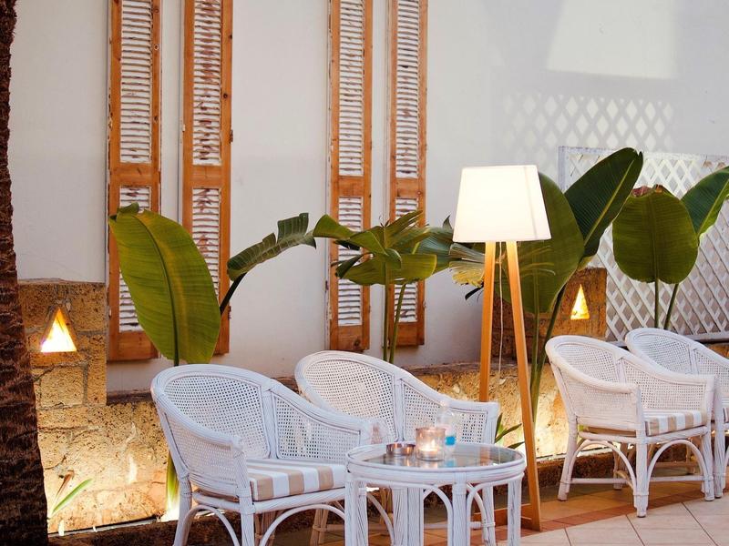 Sezení s bílými ratanovými židlemi a lampami před bílou zdí s dřevěnými okenicemi.