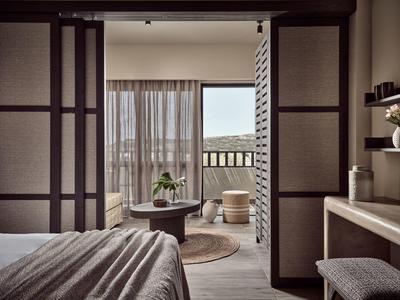 Schlafzimmer mit offener Tür zum Balkon, beiger Teppich, runde Tische, klare Vorhänge, holzfarbene Möbel.