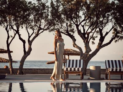 Donna in abito elegante in piedi accanto a una piscina con alberi e lettini al tramonto.