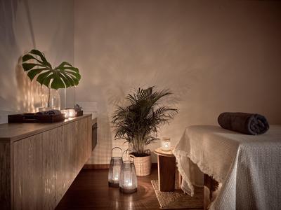 Gemütlicher Raum mit Pflanzen, Kerzenlicht, Holzmöbeln und einer mit Decke bedeckten Liege.