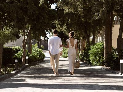 A couple walks along a shaded paved path in a garden area.