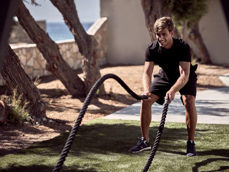 Ein Mann trainiert mit schweren Seilen auf einem grasbedeckten Weg neben Bäumen und einer Mauer.