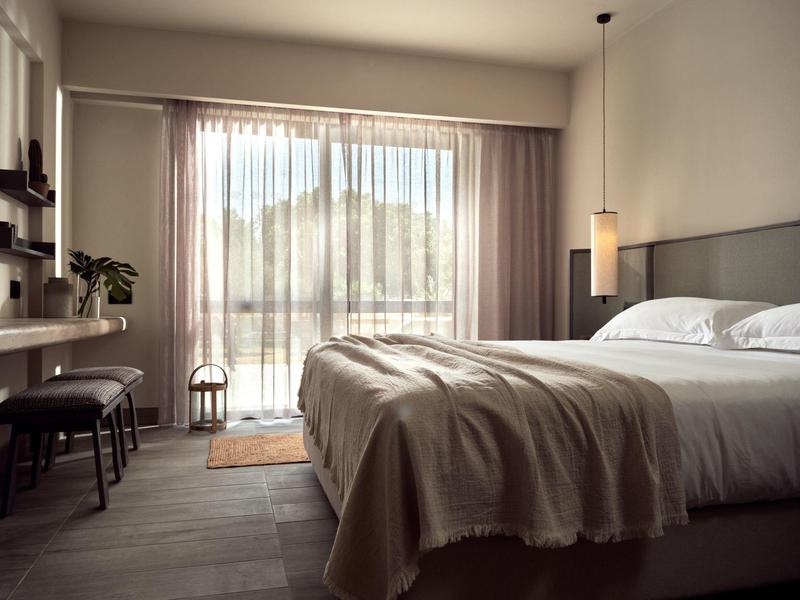 Helles Schlafzimmer mit großem Bett, Vorhängen und kleinem Schreibtisch neben Balkontür.