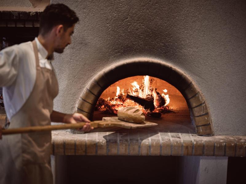 Ein Bäcker schiebt Brot mit einem Holzbrett in einen gemauerten Ofen mit offenem Feuer.