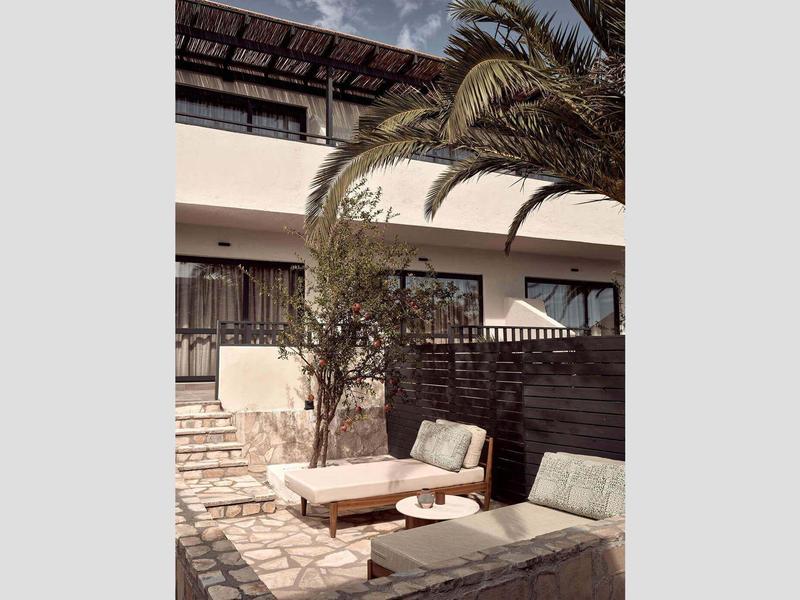 Moderner Außenbereich mit hellen Steinfliesen, Holzmöbeln und Palmen unter klarem Himmel.