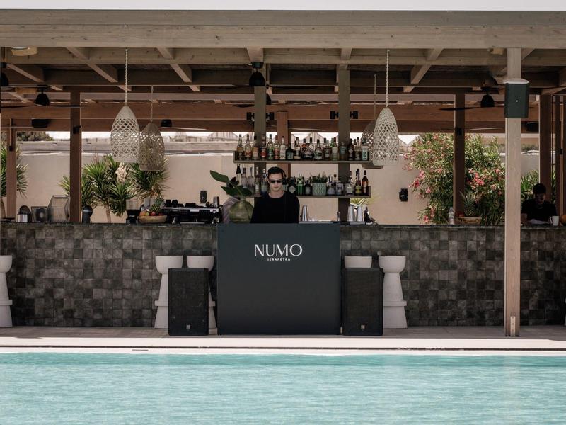Eine Person steht hinter einer schwarzen Bar mit der Aufschrift "NUMO", davor ist ein Pool.