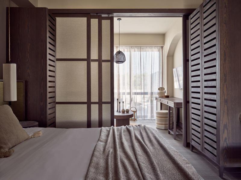 Schlafzimmer mit großem Bett, hölzernem Raumteiler und Schiebetür zum hellen Balkon mit Gardinen.