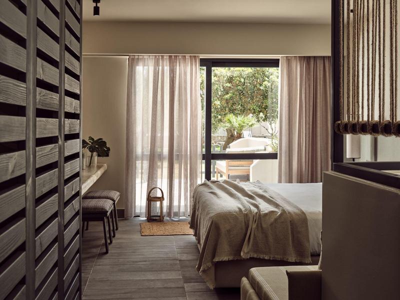 Schlicht eingerichtetes Schlafzimmer mit großem Fenster, Vorhängen und Bett mit beiger Decke.