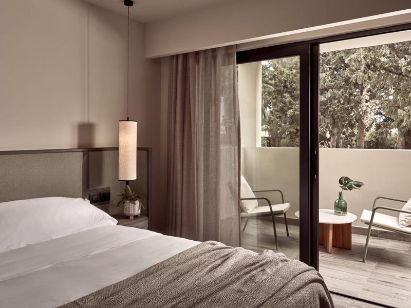 Schlafzimmer mit Doppelbett und Zugang zu Balkon mit Stühlen und kleinem Tisch, helle Farbgebung.