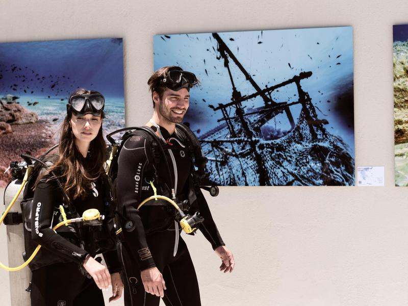 Zwei Taucher in kompletter Ausrüstung stehen vor Bildern eines Schiffswracks im Museum.