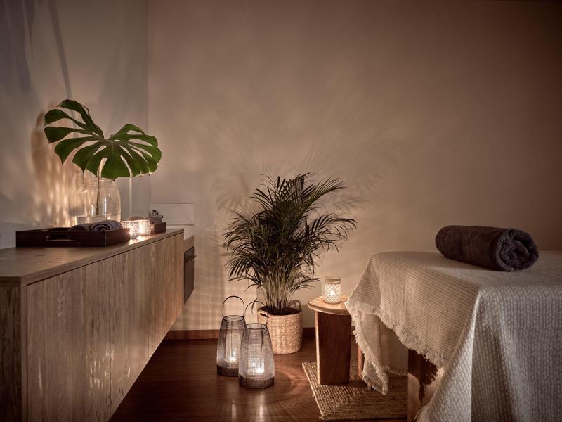 Gemütlicher Raum mit Pflanzen, Kerzenlicht, Holzmöbeln und einer mit Decke bedeckten Liege.