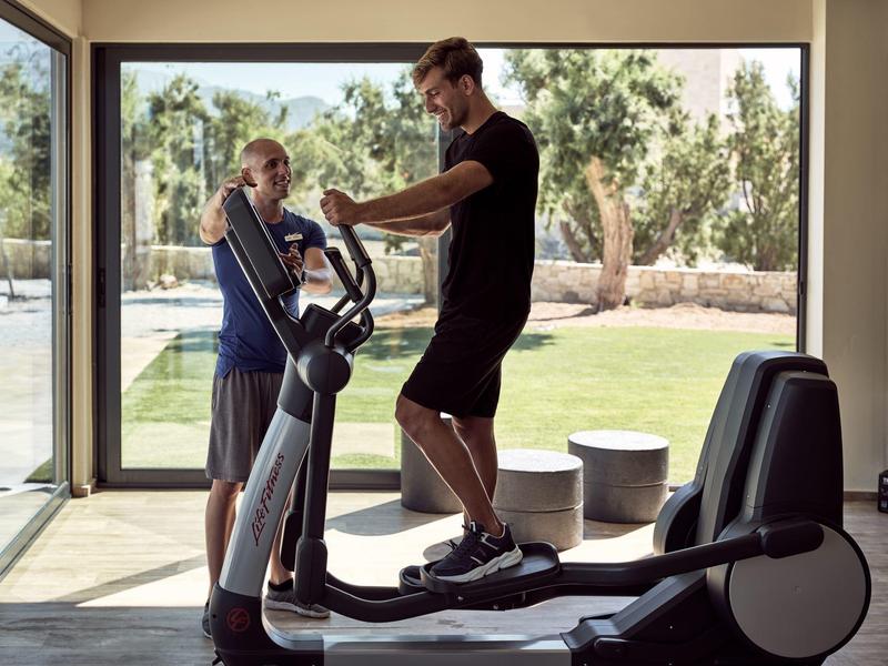 Zwei Männer trainieren auf einem Crosstrainer vor großen Fenstern mit Blick auf Garten und Pool.