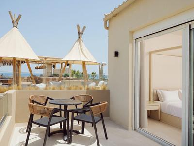 Balcony with table and chairs overlooking beach tents and sea next to a room with open sliding door.