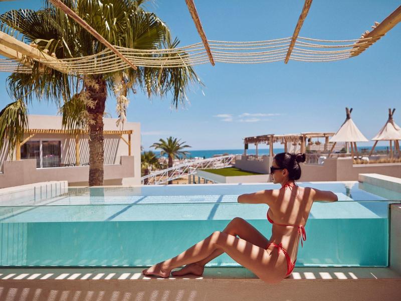 Woman in red swimsuit relaxing by pool with sea view and palm trees on a sunny day.