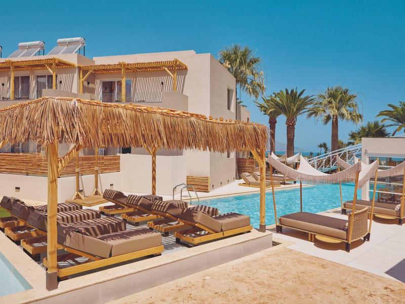 Hotel pool with loungers and straw sunshades under clear blue sky.