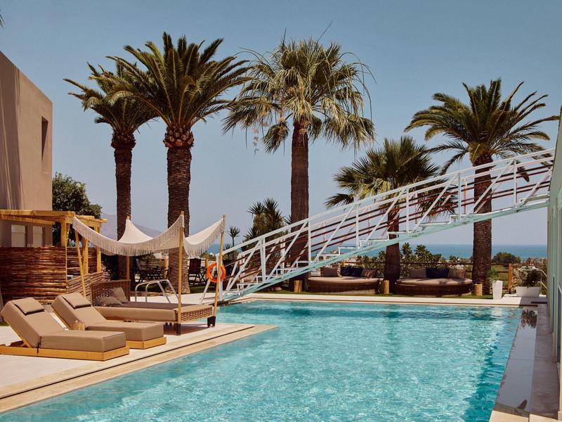 Modern pool area with sun loungers, palm trees, and sea view under clear sky.