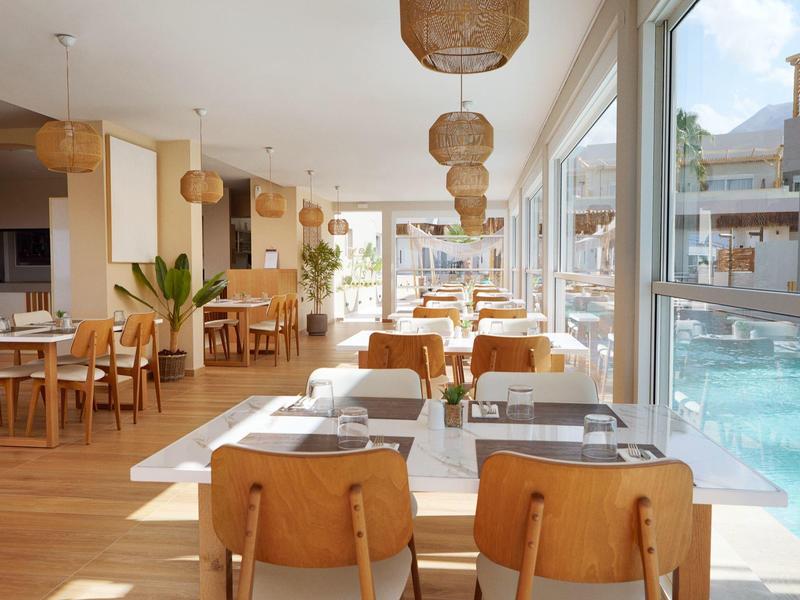 Bright, modern restaurant with wooden furniture and pool view through large windows.