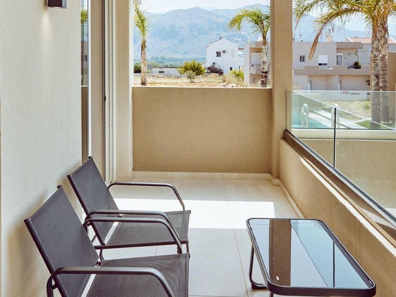 Balcony with two chairs, glass table, and view of palm trees and mountains under clear sky.