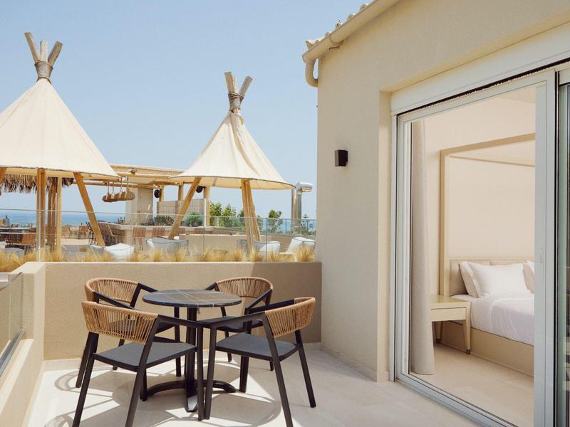 Balcony with table and chairs overlooking beach tents and sea next to a room with open sliding door.