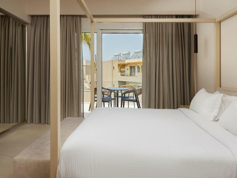 Modern hotel room with large bed and balcony featuring chairs and table.