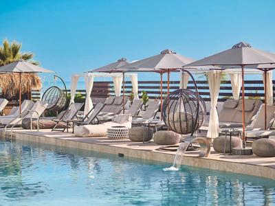 Poolbereich mit Liegestühlen, Sonnenschirmen und Hängesesseln vor blauem Himmel und Wasser.