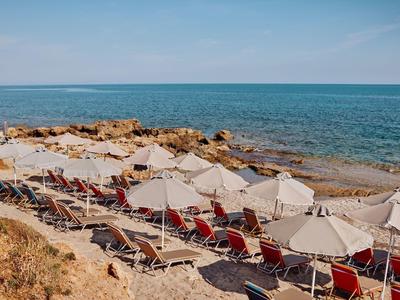 Strand mit roten Liegestühlen und weißen Sonnenschirmen, blaues Meer, felsige Küste, klarer Himmel.